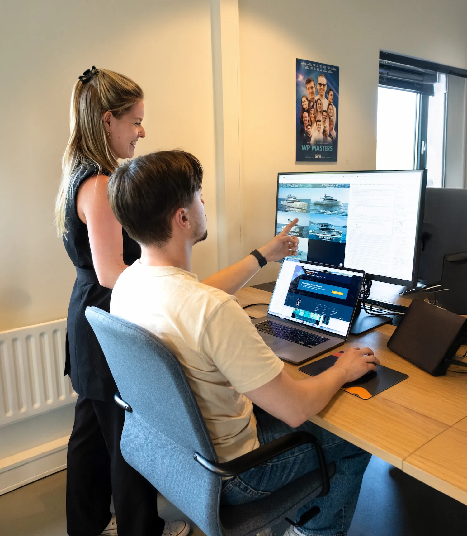 Twee collega's werken samen aan website-ontwikkeling op een kantoor, terwijl ze naar schermen met webpagina's en ferry-afbeeldingen kijken.